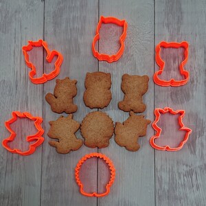 May include: An assortment of orange plastic cookie cutters in various animal shapes, including an owl, bear, and squirrel, alongside baked cookies in matching forms. The cookies are golden brown, and the cutters are arranged on a light gray wooden surface.