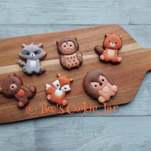 May include: A wooden cutting board displays seven woodland animal cookies: a raccoon, owl, beaver, bear, fox, and hedgehog. The cookies are brown, gray, orange, and white. The text "Bee's Cookie Jar" is visible on the board.
