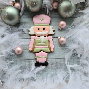 Op de afbeelding: Een gingerbread-nootkrakerkoekje versierd met roze, groene en bruine glazuur. Het koekje is omringd door witte veren en groene en roze kerstballen.