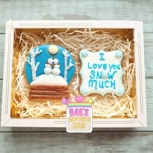 May include: A gift box filled with shredded paper, containing two decorated cookies. One cookie is a snow globe design with two snowmen, and the other says "I LOVE YOU SNOW MUCH". A jar of cookies is also visible.