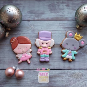 Puede incluir: Tres galletas de azúcar decoradas con personajes de El Cascanueces: una bailarina con un vestido rosa y blanco, un soldado cascanueces con un sombrero morado y un uniforme rojo y verde, y un rey ratón con un abrigo azul y blanco y una corona dorada.