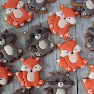 May include: An assortment of decorated cookies featuring woodland animal designs. The cookies include orange foxes and gray raccoons, each with white accents and black details. The cookies are arranged on a light blue wooden surface.