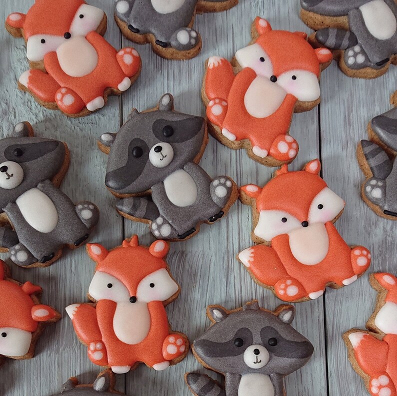 May include: An assortment of decorated cookies featuring woodland animal designs. The cookies include orange foxes and gray raccoons, each with white accents and black details. The cookies are arranged on a light blue wooden surface.