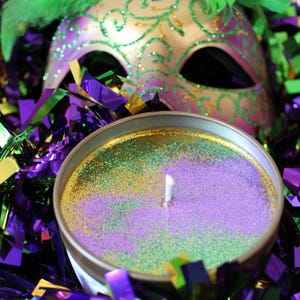 May include: A round candle in a silver tin with purple, green, and gold glitter. A white wick is centered in the candle. A decorative mask with green and purple accents and green feathers is in the background, along with purple and gold streamers.