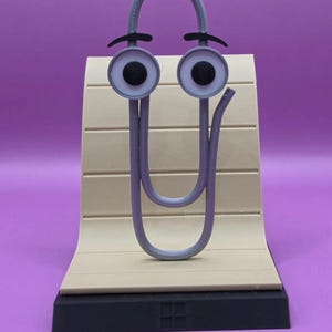 May include: A grey paperclip character figure with large white eyes, black pupils, and black eyebrows. The figure is mounted on a beige, curved, plank-like stand with horizontal lines, supported by a solid black rectangular base.