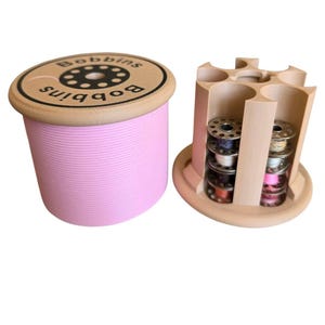 May include: A light pink cylindrical bobbin storage container with a tan lid labelled "Bobbins" and a matching tan open-frame organiser holding several metal sewing machine bobbins with various coloured threads wound around their centres.
