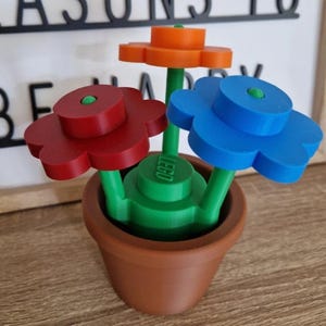 May include: A brown plastic flower pot containing three constructed plastic flowers. The flowers have red, orange, and blue petals with green stems. The central green base features the embossed text LEGO. The design consists of interlocking plastic building blocks.