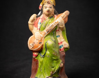 Puede incluir: Pequeña figura envejecida de una deidad, probablemente de origen indio. La figura viste una prenda verde, una cofia dorada y sostiene un instrumento de cuerda. La figura tiene una base marrón rojiza y se alza sobre un fondo oscuro.