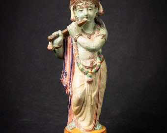Puede incluir: Una figurita pintada de verde y blanco de Krishna tocando una flauta. La figurita está de pie sobre una base marrón y tiene un tocado colorido.