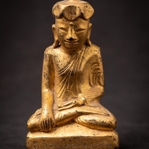 May include: A small, gold-colored statue of a seated figure in a meditative pose. The figure wears a distinctive cap and a robe draped over one shoulder. The surface shows signs of wear with dark, weathered patches. Inscribed text is visible on the base.