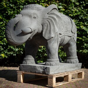 Op de afbeelding: Een groot grijs stenen olifantenbeeld met ingewikkelde houtsnijwerk. De olifant staat op een houten pallet.