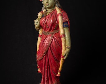 Puede incluir: Estatua de pie de una mujer con piel verde, vestida con un sari rojo y joyas doradas. La estatua tiene un tocado detallado y sostiene una flor. La base es de color marrón oscuro.