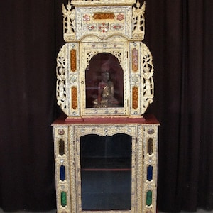 May include: A tall, ornate wooden cabinet with a gold finish and intricate carvings. The cabinet has two glass doors and a small, enclosed space at the top. The cabinet is decorated with colorful glass panels and sits on four carved animal feet.