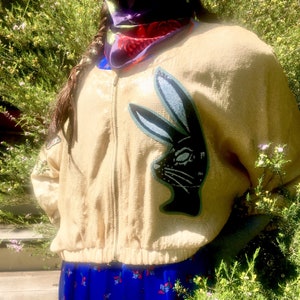 Könnte beinhalten: Beigefarbene Pailletten-Bomberjacke mit einem schwarzen und blauen Hasenmotiv auf der Vorderseite. Die Jacke hat einen Reißverschluss und gerippte Bündchen. Die Person trägt einen blauen Rock mit Blumenmuster.