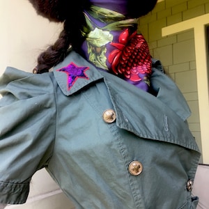 May include: A dark green short-sleeved jacket with large gold buttons. A purple sequined star is sewn on the collar. A floral scarf in shades of purple, green, and red is worn underneath the jacket.