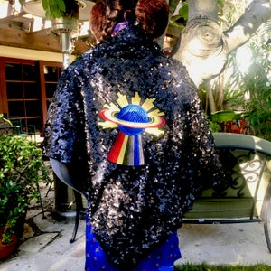 May include: A black sequined cape with a colorful embroidered emblem featuring a blue sphere with a ring and sunburst design. The cape is worn over a blue patterned garment, with a glimpse of a garden setting in the background.