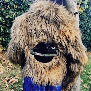 May include: A shaggy brown faux fur coat with a black sequined hat. The hat has a silver band. The coat is hanging on a tree. The bottom of a blue patterned garment is visible.