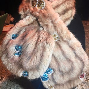 May include: A luxurious, light brown fur scarf with a striped pattern. The scarf is adorned with heart-shaped embellishments featuring gold, blue, and red sequins. The scarf is displayed on a black stand.