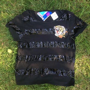 May include: Black t-shirt with horizontal sequin stripes and a tiger patch. The tiger patch is embroidered with yellow, orange, and white details. The shirt has short sleeves and a crew neck.