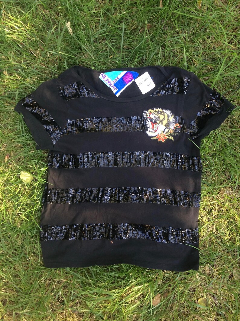May include: Black t-shirt with horizontal sequin stripes and a tiger patch. The tiger patch is embroidered with yellow, orange, and white details. The shirt has short sleeves and a crew neck.