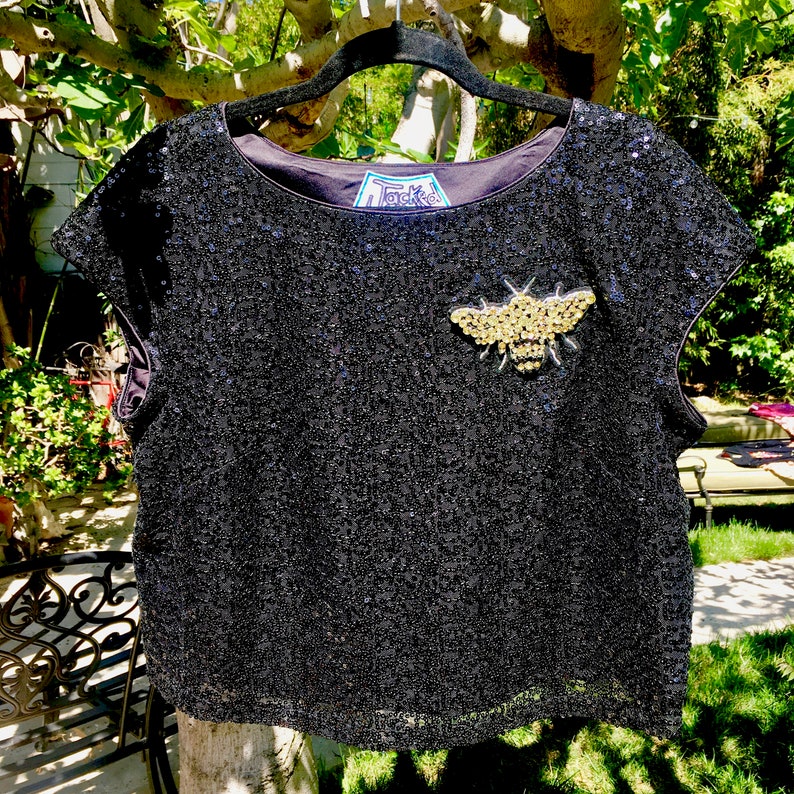May include: Black sequined top with a gold and silver bee embellishment. The top has a scoop neck and short sleeves. The label inside the top reads "Tacked". The top is hanging on a black hanger.