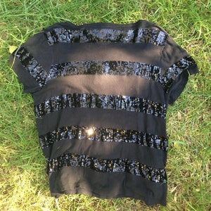 May include: Black t-shirt with horizontal bands of black sequins. The short-sleeved shirt is lying on a bed of green grass. The sequins reflect light, creating a shimmering effect. The shirt is a casual top.