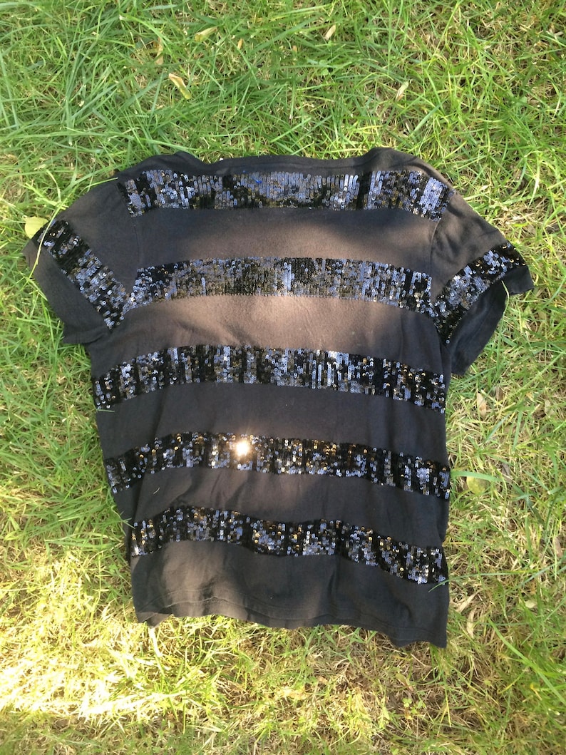 May include: Black t-shirt with horizontal bands of black sequins. The short-sleeved shirt is lying on a bed of green grass. The sequins reflect light, creating a shimmering effect. The shirt is a casual top.