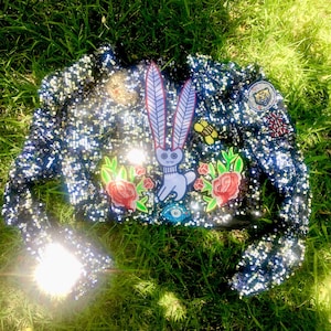 May include: A cropped, navy blue sequined jacket with various embroidered patches. The patches include a rabbit with feather-like ears, roses, and a tiger. The jacket is embellished with sequins and is lying on green grass.