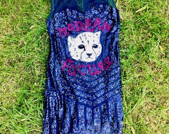 May include: A sleeveless, knee-length blue sequined dress with a sheer navy neckline. The dress features a white cheetah face with the words "MODERN KICKS" in pink sequins. The dress is lying on a bed of green grass.