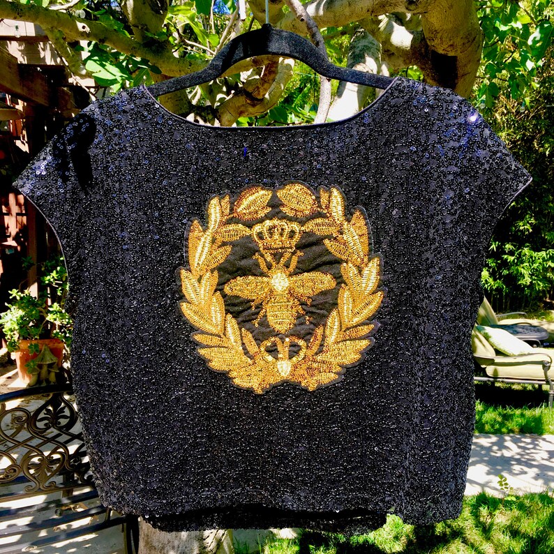 May include: A black sequined top with a round neckline. The top features a gold embroidered emblem of a bee within a laurel wreath and a crown. The garment is hanging on a black hanger against a backdrop of greenery.