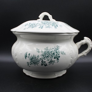 May include: White ceramic chamber pot with a lid and handle, adorned with teal floral patterns. The pot has a rounded shape with a decorative handle and a matching lid. A classic piece of antique bathroom decor.