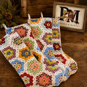 Könnte beinhalten: Zwei gesteppte Weihnachtsstrümpfe mit einem bunten Patchwork-Muster aus Stoff-Sechsecken in roten, gelben, blauen und grünen Blumen- und geometrischen Drucken. Jeder Strumpf hat eine weiße Stoffschlaufe an der Oberseite zum Aufhängen.