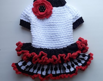May include: White crocheted dress with black and red trim. The dress features a red flower on the bodice and a piano key pattern on the skirt.