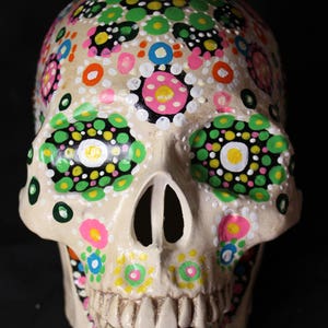 Op de afbeelding: Een witte keramische schedel geschilderd met kleurrijke stippen en bloemmotieven. De schedel heeft een gedetailleerde mond met tanden en is versierd met groene, roze, gele, oranje en blauwe verf.