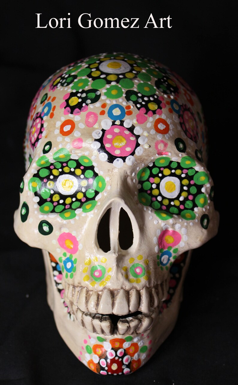 Op de afbeelding: Een witte keramische schedel geschilderd met kleurrijke stippen en bloemmotieven. De schedel heeft een gedetailleerde mond met tanden en is versierd met groene, roze, gele, oranje en blauwe verf.