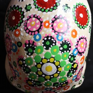 Op de afbeelding: Een witte keramische schedel versierd met kleurrijke stippen en cirkels in tinten rood, roze, groen, geel, blauw en zwart. De schedel is geschilderd op een witte achtergrond en de stippen en cirkels zijn gerangschikt in een patroon.