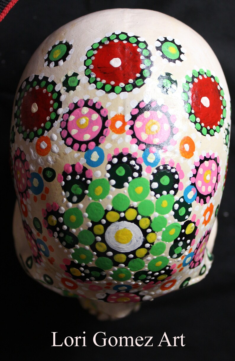 Op de afbeelding: Een witte keramische schedel versierd met kleurrijke stippen en cirkels in tinten rood, roze, groen, geel, blauw en zwart. De schedel is geschilderd op een witte achtergrond en de stippen en cirkels zijn gerangschikt in een patroon.