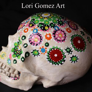 Op de afbeelding: Een witte keramische schedel versierd met kleurrijke stippen en cirkels in tinten roze, groen, rood, blauw en zwart. De schedel is geschilderd in een dot mandala-stijl.
