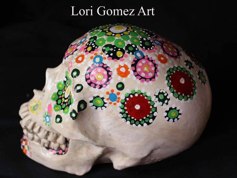 Op de afbeelding: Een witte keramische schedel versierd met kleurrijke stippen en cirkels in tinten roze, groen, rood, blauw en zwart. De schedel is geschilderd in een dot mandala-stijl.