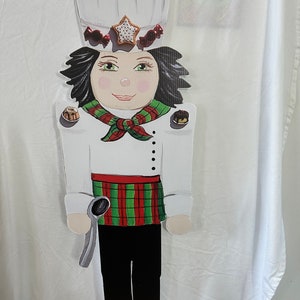 May include: A wooden cutout of a chef wearing a white chef's hat, a red and green plaid skirt, and holding a spoon. The chef is surrounded by small, colorful, painted pastries.