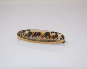 May include: An oval-shaped brooch with a gold-colored frame. The brooch features a translucent stone with black and brown patterns. The pin is a classic accessory, suitable for adding a touch of elegance to any outfit.