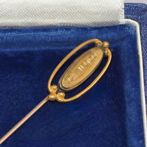 May include: A gold-toned stick pin with an oval-shaped head. The head features the word "COLLEGE" in a vertical orientation. The pin is set against a blue velvet background, suggesting a presentation box.