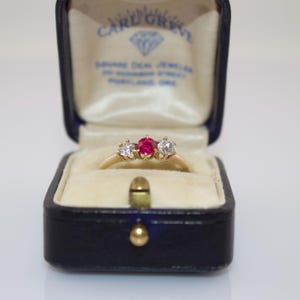 May include: A gold ring with a central red gemstone flanked by two smaller clear gemstones, displayed in a vintage blue jewelry box. The box interior is cream-colored with text that reads "CARL GREVY SQUARE DEAL JEWELS".