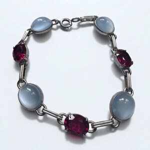 May include: A silver bracelet featuring alternating oval-shaped, light blue gemstones and oval-shaped, deep red gemstones. The bracelet is linked together with silver bars and has a clasp closure.