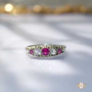May include: A gold ring featuring a five-stone setting with a central round-cut ruby flanked by two clear round-cut diamonds and two smaller outer rubies. The stones are set in a metal band with decorative prongs and a polished finish.