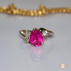 May include: A silver-toned ring featuring a prominent pear-shaped, vibrant pink gemstone at the center, flanked by two small, clear round stones on either side of the band. The metal band has a smooth, polished finish and a bypass-style design.