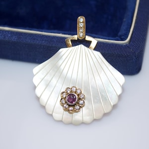 May include: A seashell-shaped pendant with a gold bail and a gold flower with a purple center stone and surrounding clear stones. The pendant is white with a pearlescent sheen and is set against a blue velvet background.