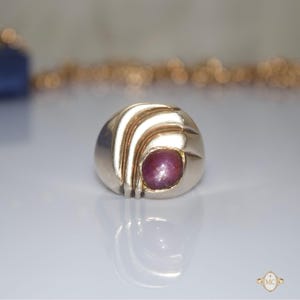 May include: A gold ring featuring a smooth, polished band with three raised, curved lines on one side and a single, round, cabochon-cut deep pink star ruby gemstone set securely into the metal surface, creating a bold and solid appearance.