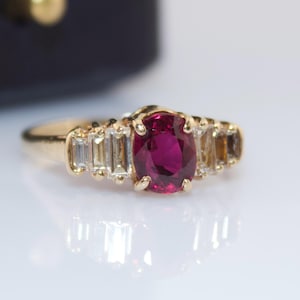 May include: A gold ring featuring a central oval-cut deep pink ruby gemstone held in a four-prong setting. The band is accented on each side by three clear baguette-cut diamonds arranged in a graduated step pattern along the gold shank.