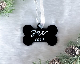 May include: Black bone-shaped Christmas ornament with the name "Jax" and the year "2023" written in white on it. The ornament is hanging from a silver ribbon.
